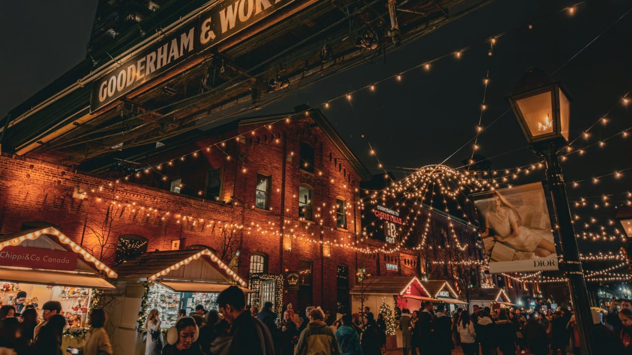 An image of downtown Toronto’s distillery district at night in the winter. There are string lights lit up and people walking around looking at shops.